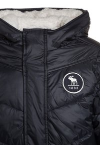 Sort polstret jakke med en hætte foret med kunstpels, diamantquiltet tekstur og et rundt logo med en elg, grundlagt 1892.