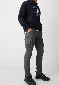 Szare spodnie cargo z wieloma bocznymi kieszeniami i slim fit, w połączeniu z granatowym hoodie z białym wzorem graficznym. Czarne buty.