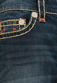 Tela de denim oscura con costuras amarillas contrastantes y pequeños acentos rojos. Presenta remaches de metal y un acabado texturizado.