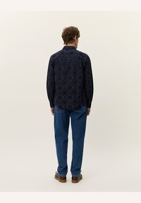 Marineblå langermet skjorte med geometrisk mønster og teksturert stoff, kombinert med lyseblå jeans og brune sko. Visning fra baksiden.