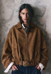 Tânără cu păr lung și închis la culoare, purtând o jachetă supradimensionată din piele întoarsă maro cu curea și manșete albe la cămașă, stând în fața unui zid texturat.