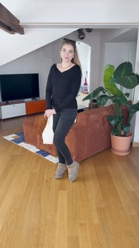 Black long-sleeve top, dark pants, and gray slippers. Light wood floor, brown corduroy couch, green plant, and modern TV stand in a bright space.