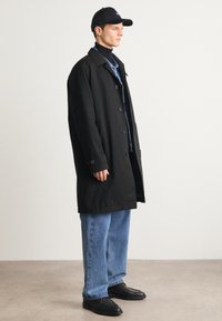Manteau noir avec fermeture à boutons, porté sur une chemise en denim et un jean bleu. Une casquette noire et des chaussures noires complètent le look. Design minimaliste.