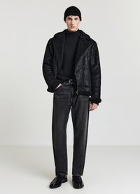 Giacca nera foderata in shearling con chiusura a zip, abbinata a jeans grigio scuro e scarpe nere lucide. Il modello indossa un berretto nero in maglia.