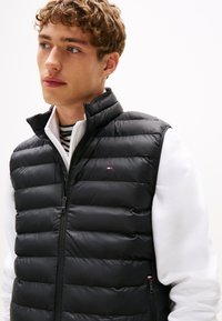 Jovem com cabelo encaracolado vestindo um colete preto acolchoado sobre uma jaqueta branca com uma camisa às riscas por baixo, olhando para a esquerda.