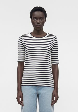 Γυναίκα με κοντά σγουρά μαλλιά που φοράει μια ασπρόμαυρη ριγέ μπλούζα με μανίκια μέχρι τους αγκώνες και ανοιχτό μπλε τζιν μπροστά σε απλό φόντο.