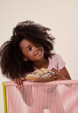Niña joven con cabello rizado sonríe mientras sostiene un zapato beige con estampado de leopardo, apoyada en una silla rosa tejida.
