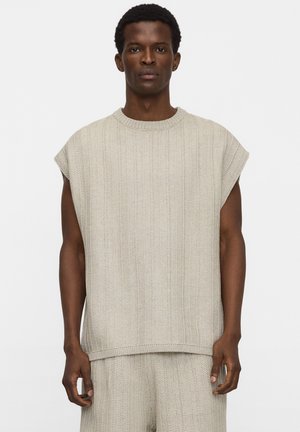 Homme portant un pull sans manches beige en tricot et un pantalon assorti en tricot, debout face à l'avant sur un fond uni.
