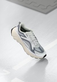 Buty sportowe z siateczkową cholewką w odcieniach szaro-białych, z teksturowanym wzorem, amortyzowaną podeszwą oraz trwałą czarną podeszwą zewnętrzną.