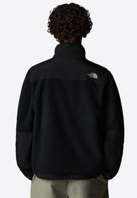 Black fleece jacket with a high collar, contrasting black nylon shoulder and sleeve panels, and a logo on the upper back. Textured fabric.