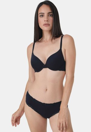 Frau mit langen dunklen Haaren, die einen schwarzen gepolsterten BH und passende schwarze Unterwäsche mit Spitzenbesatz vor einem schlichten Hintergrund trägt.