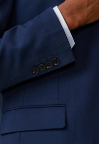 Marineblauer Blazer aus strukturiertem Stoff, mit einer einzelnen Tasche und vier schwarzen Knöpfen am Ärmelsaum, dazu ist ein weißer Hemdkragen sichtbar.
