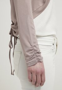 Jasnobrązowa bluzka z długim rękawem, zebrana w akcentach na rękawach, w połączeniu z białymi dopasowanymi jeansami. Bluzka ma teksturowany panel z przodu.