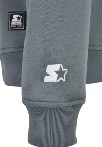 Manche en tissu gris avec poignet côtelé, ornée du logo Starter brodé en blanc et d’un patch rectangulaire noir Starter Black Label.