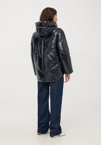 Giacca nera imbottita con finitura lucida, grande cappuccio e design a zig-zag trapuntato. Abbinata a pantaloni in denim scuro e sneakers bianche.