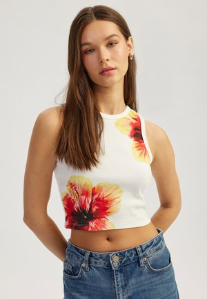 White cropped tank top with a ribbed texture, featuring vibrant red and yellow hibiscus flower prints. Paired with blue high-waisted jeans.