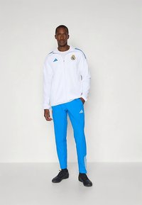 Witte trainingsjas met blauwe accenten en logo, gecombineerd met felblauwe broek met drie strepen en logo. Zwarte sportschoenen.