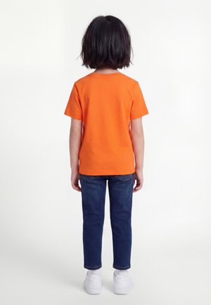 Barn med kort mørkt hår står med ryggen til, iført oransje kortermet skjorte, blå jeans og hvite joggesko på hvit bakgrunn.