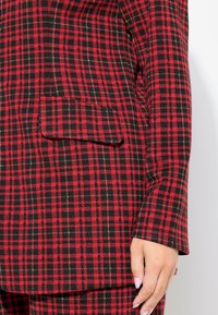 Blazer a cuadros rojo y negro con un gran bolsillo de parche, hombros estructurados y una tela suave con acentos metálicos.