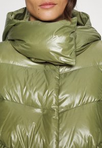 Gros plan sur une personne portant une doudoune brillante vert olive avec un col haut et rembourré couvrant le cou.