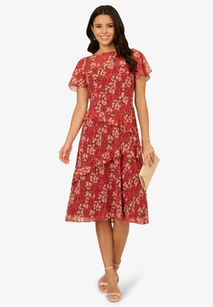 Rode bloemenjurk met gelaagde ruffles, korte flapmouwen en een getailleerde taille. Gepaard met een gouden clutch en naakte strappy sandalen.