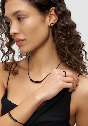 Schwarzes minimalistisches Schmuckset mit einem gebogenen Halsband, Armband und Ring mit einer strukturierten Oberfläche, getragen von einem Modell mit lockigem Haar.