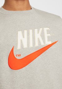 Grå sweatshirts med fet, krämfärgad och orange broderad "NIKE"-text och swoosh-logotyp på framsidan. Mjuk, texturerad tyg. Cirkulär halsringning.
