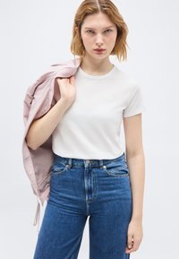 Νεαρή γυναίκα που φοράει λευκό μπλουζάκι και μπλε τζιν κρατώντας ένα ροζ μπουφάν στον ώμο της, σε φόντο απλό.