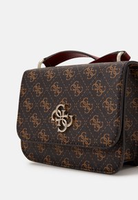 Brown patterned handbag with gold interlocking logo clasp and dark red top handle against a plain white background.