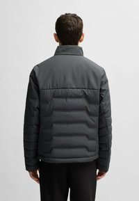 Grå pufferjacka med hög krage, vertikal quiltad mönster och elastiska ärmslut. Materialet verkar slätt och vattentåligt.