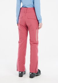 Pink ski pants with a high waist, featuring zippered pockets and side vents. Made of smooth material, paired with black ski boots.