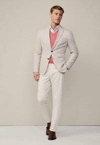 Man wearing beige blazer, pink V-neck sweater, white dress pants, and brown tassel loafers, standing against plain beige background.