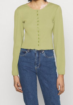 Personne portant une chemise boutonnée à manches longues vert clair côtelée et un jean bleu taille haute, debout les bras détendus le long du corps.