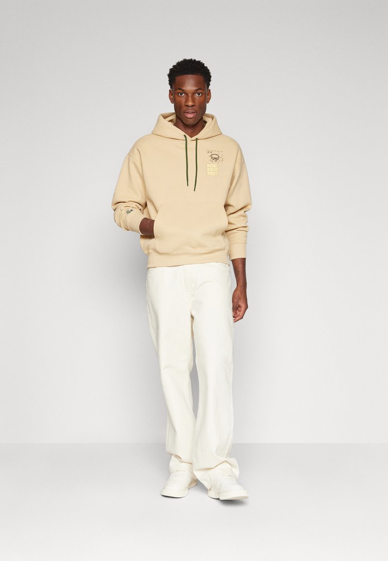Beige hoodie met groene trekkoord en grafische print op de borst, gecombineerd met witte wijde broek en witte sneakers. Soepele stoftextuur.