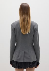 Grijze op maat gemaakte blazer met gestructureerde schouders en een rugventilatie, gecombineerd met een zwarte, volumineuze rok. De stof lijkt glad met een lichte glans.