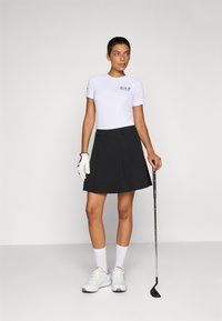 Camicia bianca a maniche corte con logo EA7, abbinata a una gonna plissettata nera e scarpe da ginnastica bianche, mentre tiene una mazza da golf in mano guantata.