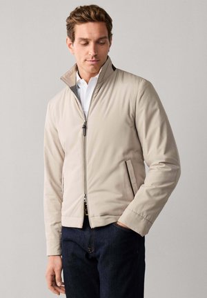 Man die een beige jas met ritssluiting draagt over een wit overhemd en donkere spijkerbroek, staand tegen een effen lichtgrijze achtergrond met één hand in de zak.