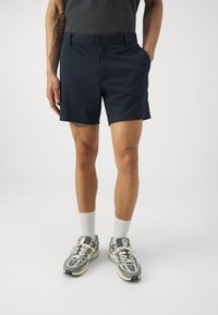 Navy shorts with a flat front, waistband button, two side pockets, and a mid-thigh length; paired with gray sneakers and white socks.