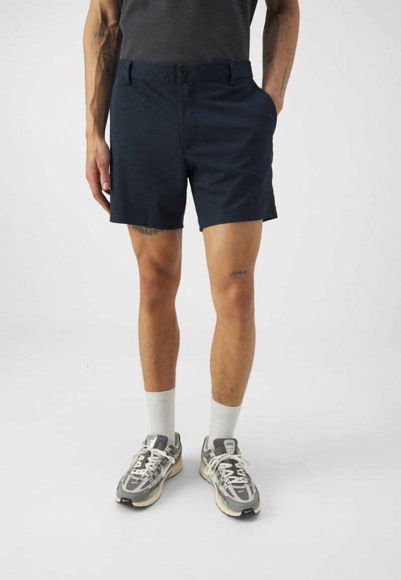 Navy shorts with a flat front, waistband button, two side pockets, and a mid-thigh length; paired with gray sneakers and white socks.