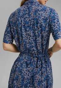Robe fleurie bleue en tissu doux, avec manches courtes, taille nouée et col arrière, ornée de motifs floraux roses et bleus.