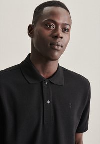 Zwart poloshirt van gestructureerde stof, met een kraag, twee knopen en een klein geborduurd logo op de borst.