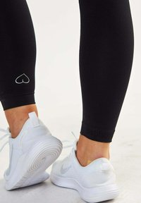Leggings neri con un piccolo logo a forma di cuore bianco sul polpaccio, abbinati a scarpe da ginnastica bianche con dettagli grigi e suole testurizzate.