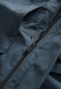Chaqueta azul marino con una textura suave y mate, que cuenta con una cremallera central negra y cierres a presión, además de un detalle de bolsillo.