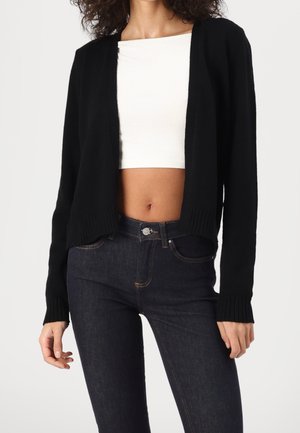 Cardigan noir en tricot avec une ouverture sur le devant, ourlet et poignets côtelés, porté sur un crop top blanc, associé à un jean en denim foncé.