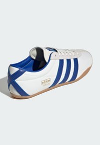 Vit sneaker med blå accenter, med tre horisontella ränder, mocka tåhuvud och en guldig logotyp. Gummisula med strukturerad detalj.