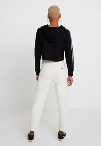 Černá zkrácená mikina s kapucí a bílými pruhy na rukávech, spárovaná s bílými širokými džíny a černými teniskami. Na džínách jednoduché trojúhelníkové logo.