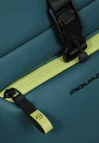 Türkise wasserdichte Tasche mit strukturierter Oberfläche, ausgestattet mit einer schwarzen Schnalle, einem neon-gelben Reißverschlussakzent und geprägtem Markenlogo.