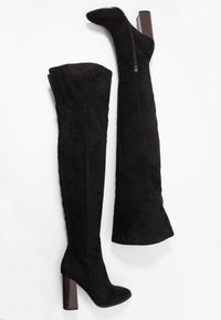 Botas negras de ante por encima de la rodilla con un tacón grueso y una cremallera lateral. Textura suave y un diseño ajustado que se extiende por encima de la rodilla.