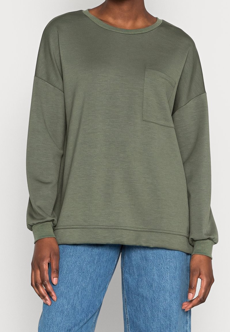 Osoba ubrana w oliwkową koszulę z długim rękawem z kieszonką na piersi oraz niebieskie dżinsy, stojąca na jednolitym tle.