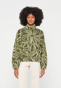 Chaqueta de forro polar verde con patrones florales blancos, cuello alto, cierre con cremallera, dos bolsillos delanteros y puños acanalados. Tejido suave y texturizado.